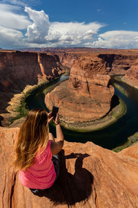 Rear view of woman using phone while sitting on cliff against sky