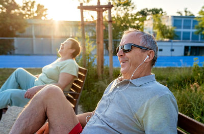 Happy smiling senior couple with earphones exercising using outdoor training exercise bike machine 