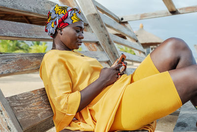 Ghana woman with african colorful headdress sitting on a bench in the village of keta with cellphone