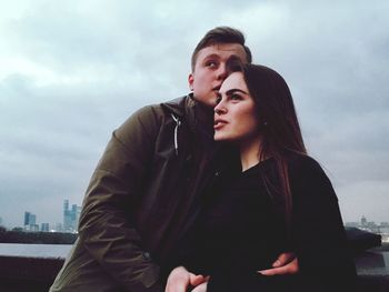 Couple looking away while standing by retaining wall against cloudy sky