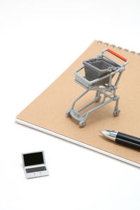 High angle view of toy laptop by shopping cart and spiral notebook against white background