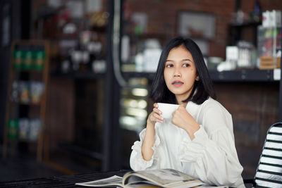 Young beautiful asian woman in white blazer with coffee cup sitting in cafe dark brown background