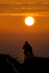 Silhouette man standing on shore against orange sunset sky