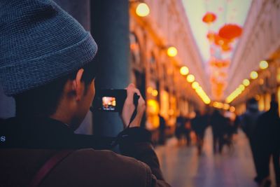 Rear view of man photographing on illuminated footpath in city