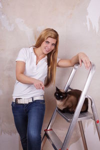Portrait of smiling beautiful woman with siamese cat on ladder against wall