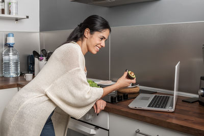 Young woman holding avocado while video conferencing over laptop at home