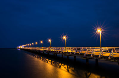 Illuminated street by sea against sky at night