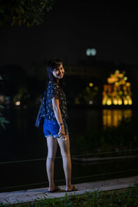 Portrait of smiling young woman standing on footpath at night