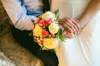 Midsection of newlywed couple holding bouquet while sitting on sofa