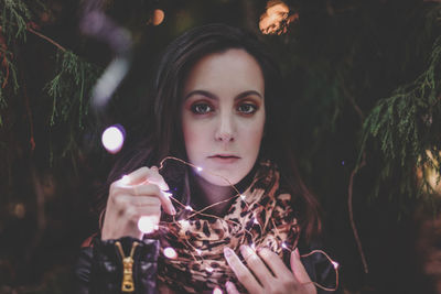 Portrait of young woman holding illuminated string lights by trees at park