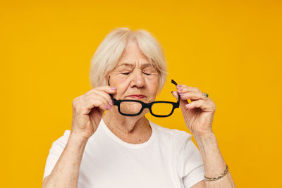 Senior woman holding eyeglasses against yellow background