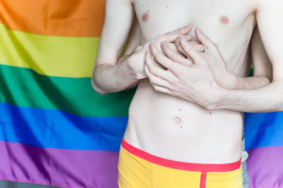 Midsection of shirtless gay couple standing against rainbow flag