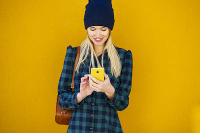 Smiling young woman using mobile phone against yellow wall