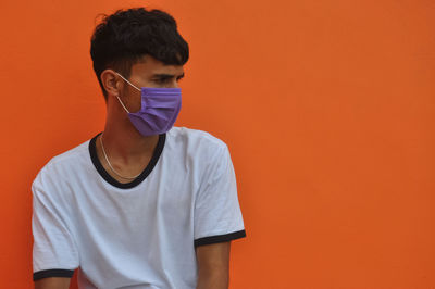 A indian young guy wearing face mask, looking sideways sitting against orange wall with copy space 