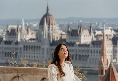 Portrait of beautiful young woman on balcony overlooking hungarian parliament in budapest, hungary