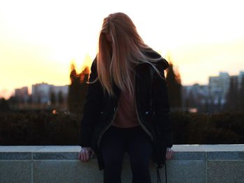 Young woman sitting on retaining wall against sky during sunset