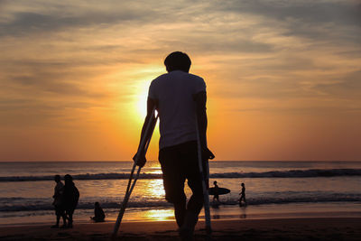 Silhouette man standing with crutches on beach against sky during sunset
