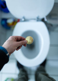 Low section of man holding bitcoin over toilet bowl while standing in bathroom