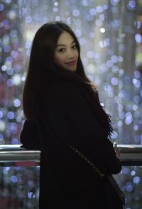 Portrait of smiling young woman standing against illuminated wall at night