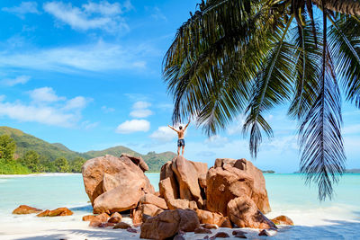 Young man with outstretched hands on rocks over tropical sandy beach.