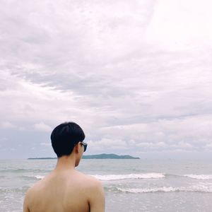 Rear view of shirtless young man standing by sea against cloudy sky