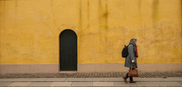 Woman carrying backpack walking on footpath by yellow wall