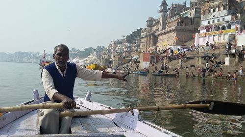 Man sitting on rowboat in river against buildings