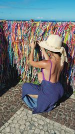 Seductive woman wearing hat tying ribbon on railing during sunny day