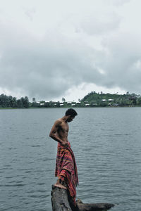 Side view of shirtless mature man with towel standing on rock in lake against cloudy sky