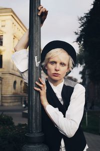 Portrait of young woman standing by pole against trees