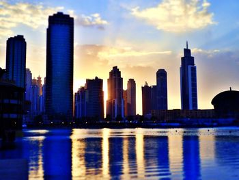 Modern buildings by river against sky during sunset