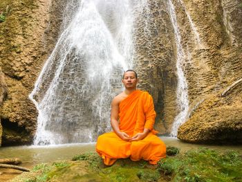 Monk meditating while sitting against waterfall