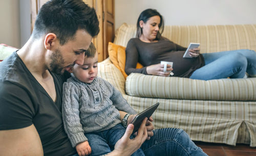 Mother holding mobile phone while father with son using digital tablet in living room