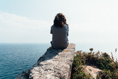 Rear view of man sitting on rock by sea against sky