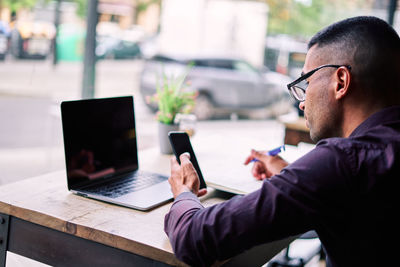 Side view of handsome young hispanic male entrepreneur checking information on smartphone and writing notes in planner while working at table with laptop