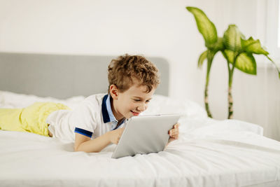 Curly-haired boy is lying on the bed in the bedroom and playing computer games on a tablet