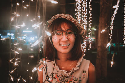 Portrait of young woman wearing eyeglasses amidst illuminated lights at night