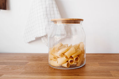 Dry raw large macaroni from a glass large jar with a wooden eco lid on a wooden countertop 