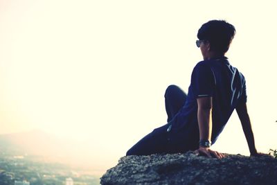 Rear view of man sitting on rock against clear sky