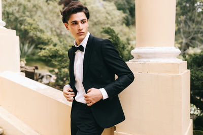 A beautiful young man, the groom in an elegant wedding suit, stands posing in the city's old park