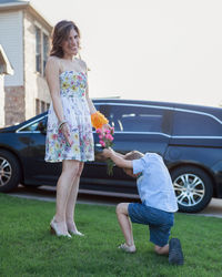 Son proposing mother while kneeling on field against clear sky