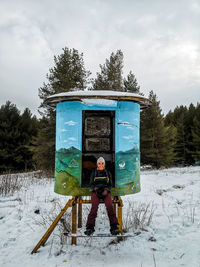 Woman sitting at painted storage tank over field against sky during winter