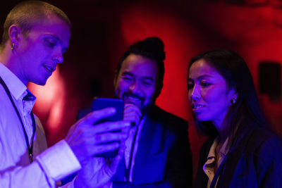 Businessman sharing mobile phone with male and female colleagues in illuminated workshop