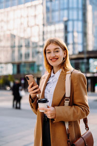 The woman in a blazer is smiling, holding a cup of coffee and a cell phone