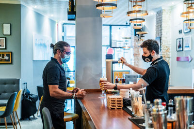 Side view of male customer in medical mask standing at counter and waiting for glass of beer served by barman