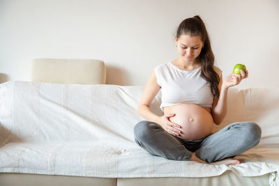 Young pregnant woman holding fruit while sitting on sofa at home