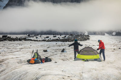 Two men set up a tent below a dramatic mountain peak.