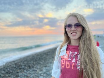 Portrait of young woman standing at beach against sky during sunset