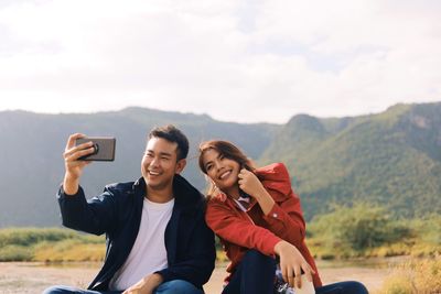 Happy couple taking selfie with mobile phone while sitting against mountains