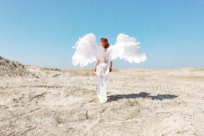 Full length of angel woman with white wings standing on sand against sky